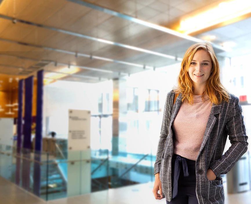 Female student with red hair, grey coat and pink blouse in front of a blurred background of Stern's lobby floor