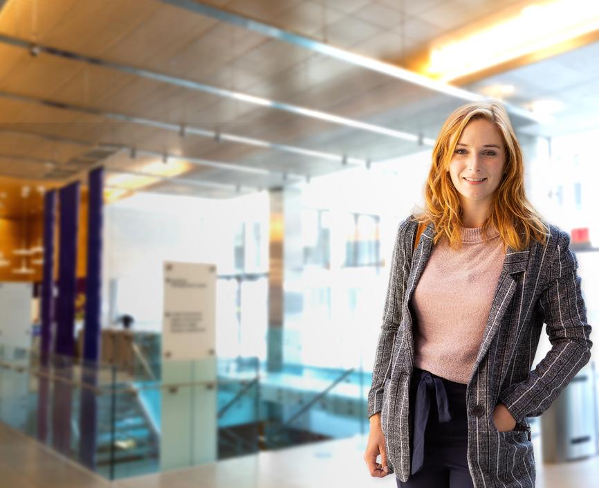 Female student with red hair, grey coat and pink blouse in front of a blurred background of Stern's lobby floor