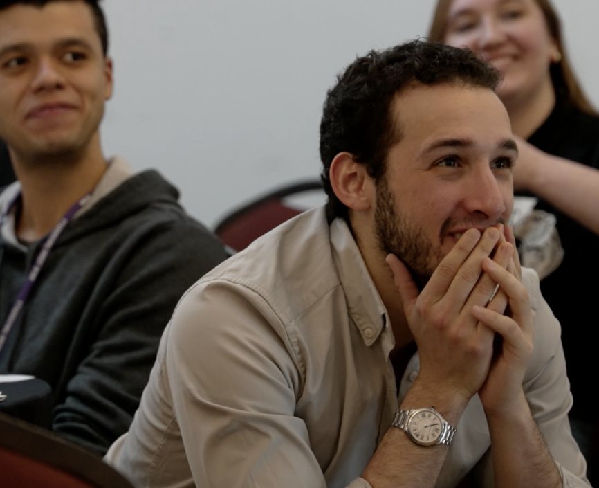 A masculine presenting person with short curly hair sits in a crowd of people, hands covering his smile in excitement and attentive listening. 