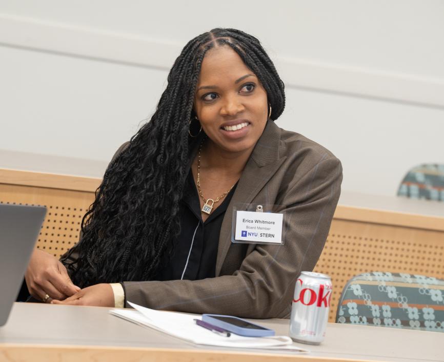 A feminine-presenting person with black hair and brown skin smiles, intrigued, as she leans forward on a desk.