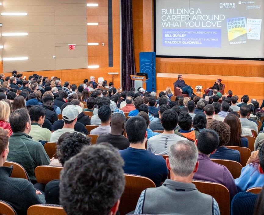 Audience in a large auditorium listening to two men on a stage.