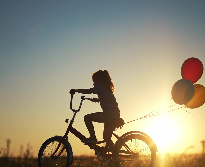In the sunset, a child rides a bike with balloons tied to the seat 