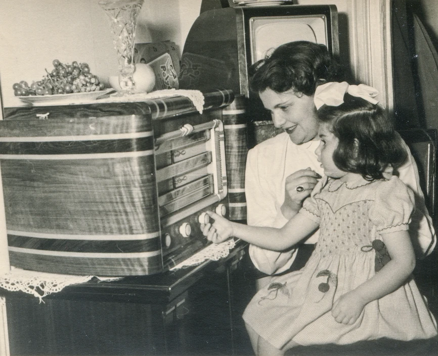 Black and white photo of a girl sitting on a woman's lap adjusting the knob on a large radio