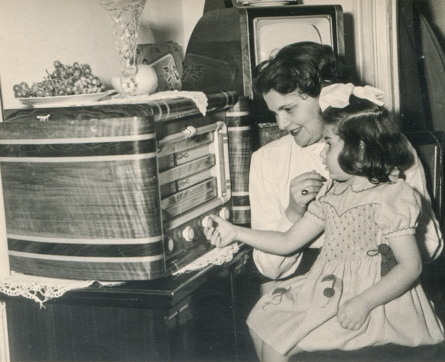 Black and white photo of a girl sitting on a woman's lap adjusting the knob on a large radio