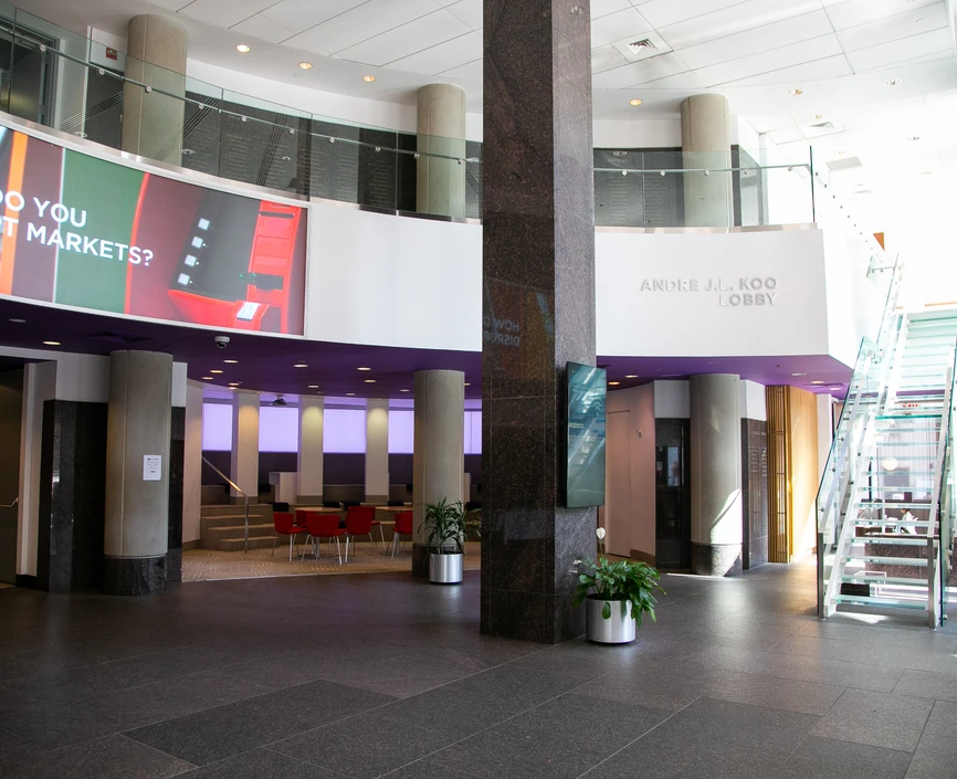 Ground floor view of empty lobby, clear stairs going up to a second level, column in the center and large screen in upper leff