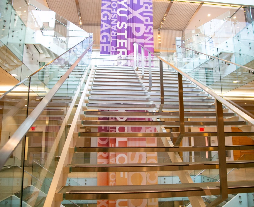 View from the bottom of a modern-looking glass stairway with a multi-colored banner behind it, located in Tisch Hall. 