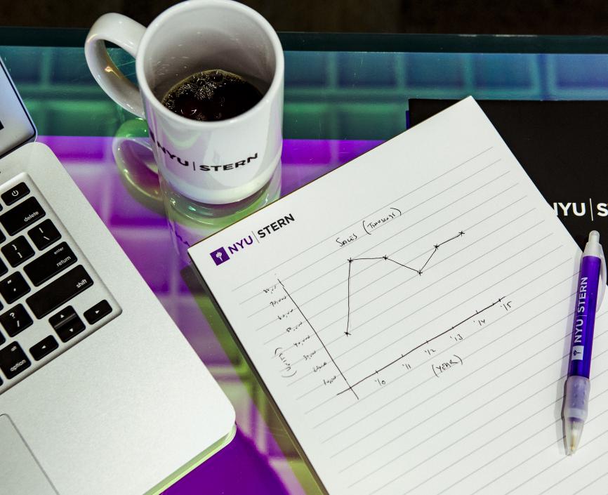 A laptop, notebook with a pen and a mug on a desk