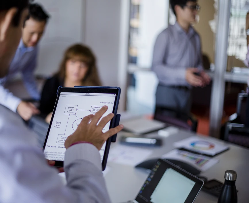Image of a small meeting, we see over the shoulder of a man holding a tablet, and in the background are 3 other employees. 