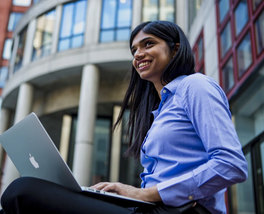Smiling young woman sitting in front of Stern building with a laptop on her lap