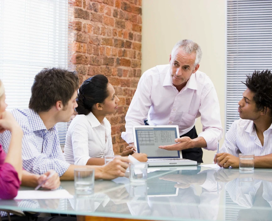 four people sitting at a glass table looking at a fifth person who is explaining something from a laptop
