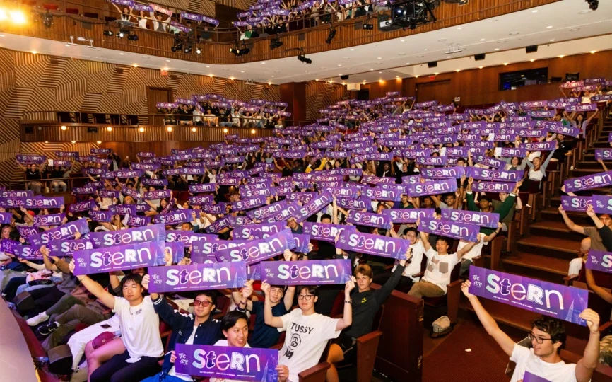 A photo of a big crowd of NYU Stern Undergraduate Students sitting in an auditorium and holding signs that say "Stern."