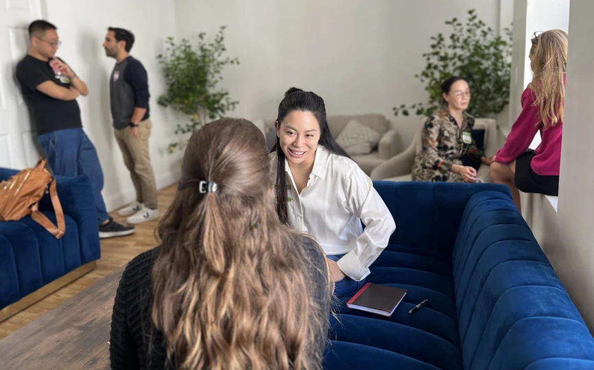 Three pairs of students sit engaged in conversation in a room featuring two large blue couches, two comfy linen chairs, and two large house plants. On the far left of the photo, two masculine-presenting students lean against the doorframe as they talk. In the center forefront, two feminine-presenting people sit on a blue couch, one with her back to the camera and the other, smiling as she looks at her partner, facing the camera. On the far right, two feminine-presenting converse in the windowsill.