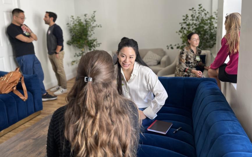 Three pairs of students sit engaged in conversation in a room featuring two large blue couches, two comfy linen chairs, and two large house plants. On the far left of the photo, two masculine-presenting students lean against the doorframe as they talk. In the center forefront, two feminine-presenting people sit on a blue couch, one with her back to the camera and the other, smiling as she looks at her partner, facing the camera. On the far right, two feminine-presenting converse in the windowsill.