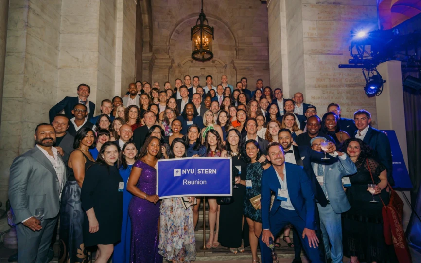A group of NYU Stern Alumni holding a flag that says "NYU Stern Reunion" celebrate at Reunion 2025.