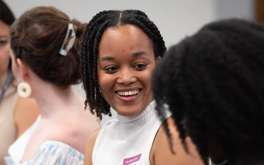 A feminine-presenting person with brown skin and dark tight curly hair smiles at the person seated next to hear,