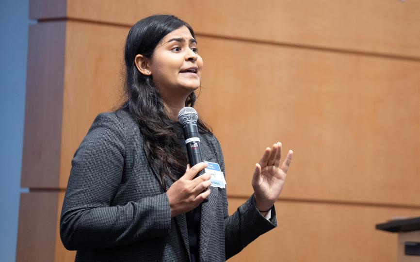 A feminine-presenting person with medium dark skin and dark curly hair tells a story with a mic in her hand.