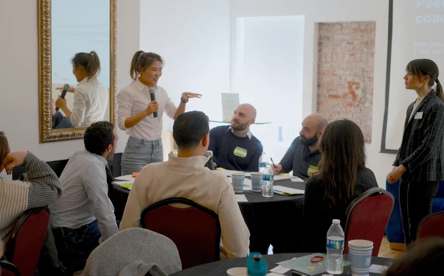 A feminin-presenting person with medium skin tone and dark hair pulled into a ponytail smiles as she speaks into a mic in a room full of peers and her professor.