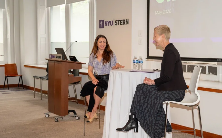 Michelle Bacharach laughs with Beth Briggs sitting at a high top table with the NYU Stern logo visible behind them