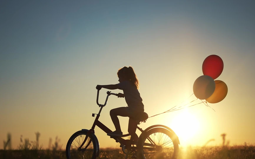 In the sunset, a child rides a bike with balloons tied to the seat 