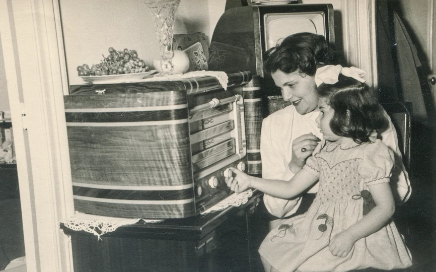 Black and white photo of a girl sitting on a woman's lap adjusting the knob on a large radio