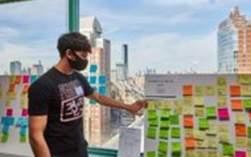 Student participating in the Design Sprint pointing to sticky notes with Brooklyn Bridge outside the window