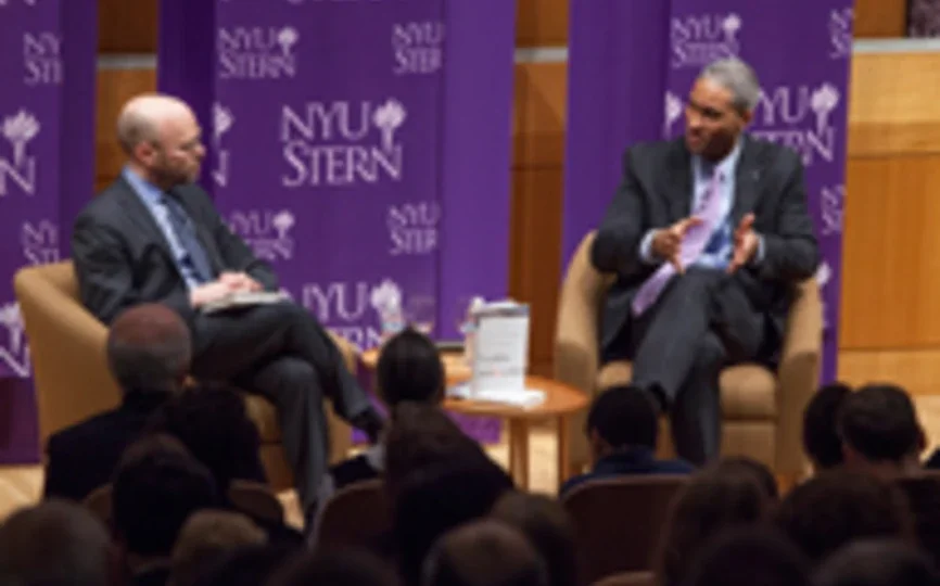 peter henry on a chair in conversation with another person with nyu stern purple banners in background