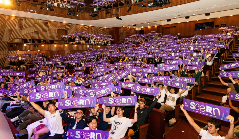 A photo of a big crowd of NYU Stern Undergraduate Students sitting in an auditorium and holding signs that say "Stern."