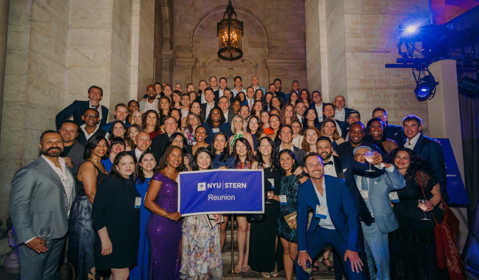 A group of NYU Stern Alumni holding a flag that says "NYU Stern Reunion" celebrate at Reunion 2025.