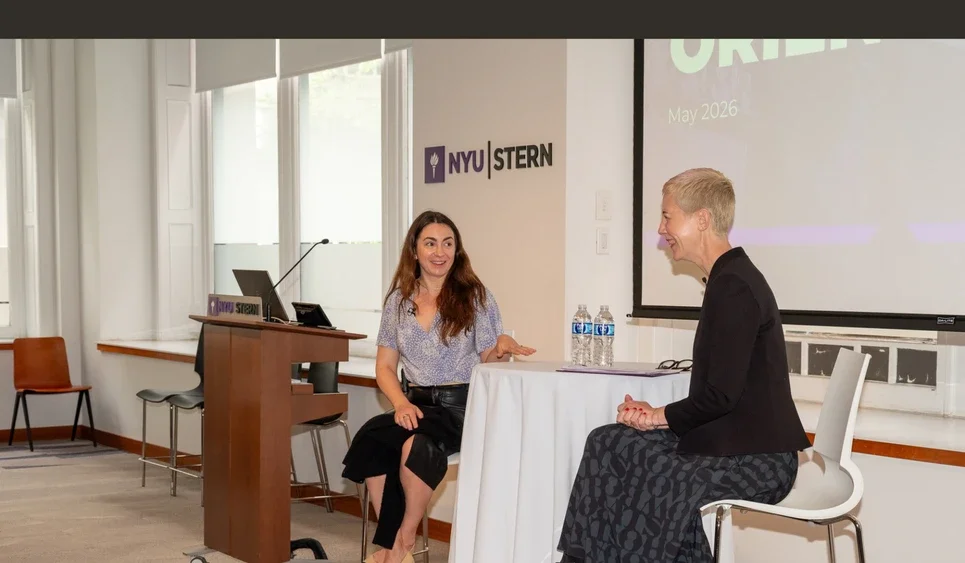 Michelle Bacharach laughs with Beth Briggs sitting at a high top table with the NYU Stern logo visible behind them