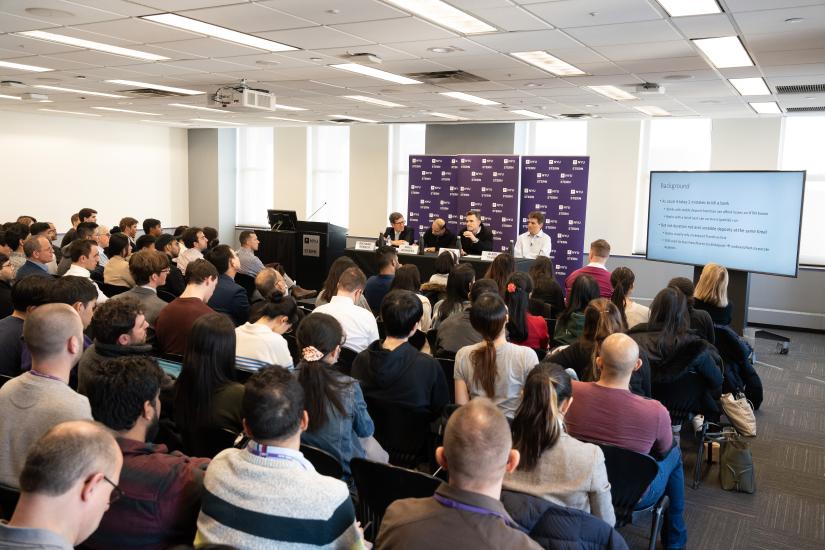 group of people watching presentation and listening to four professors speak at the front of a room