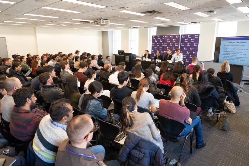 group of people watching presentation and listening to four professors speak at the front of a room