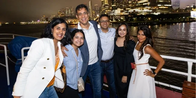 A group of 6 people stand outside at night on the deck of a boat with the NYC skyline in the background