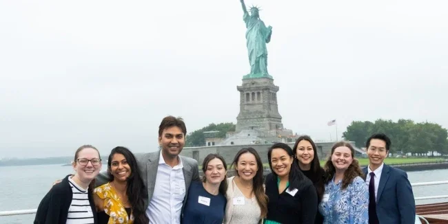 A group of 9 people stand on the deck of a boat with the Statue of Liberty visible behind them