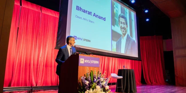 Dean Anand stands at a podium speaking to MBA students